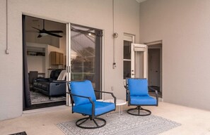 Un patio con dos sillas azules y una mesa pequeña, una puerta corrediza de vidrio que da a una sala de estar y un ventilador de techo.
