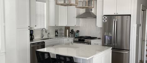 Una cocina moderna con armarios blancos, una isla central con taburetes de bar, electrodomésticos de acero inoxidable e iluminación colgante.