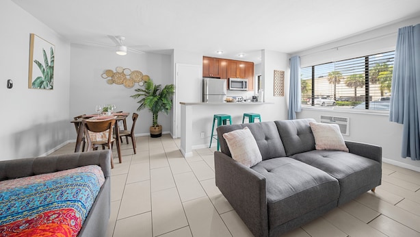 A modern living room with a grey sofa, a colorful bed, a dining area with wooden chairs, and a kitchen with wooden cabinets and a microwave. - Bayview Apartments by Lowkl (Fort Lauderdale)