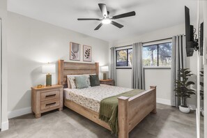 A bedroom with a wooden bed, two bedside tables with lamps, a ceiling fan, a window with curtains, and a wall-mounted TV.
