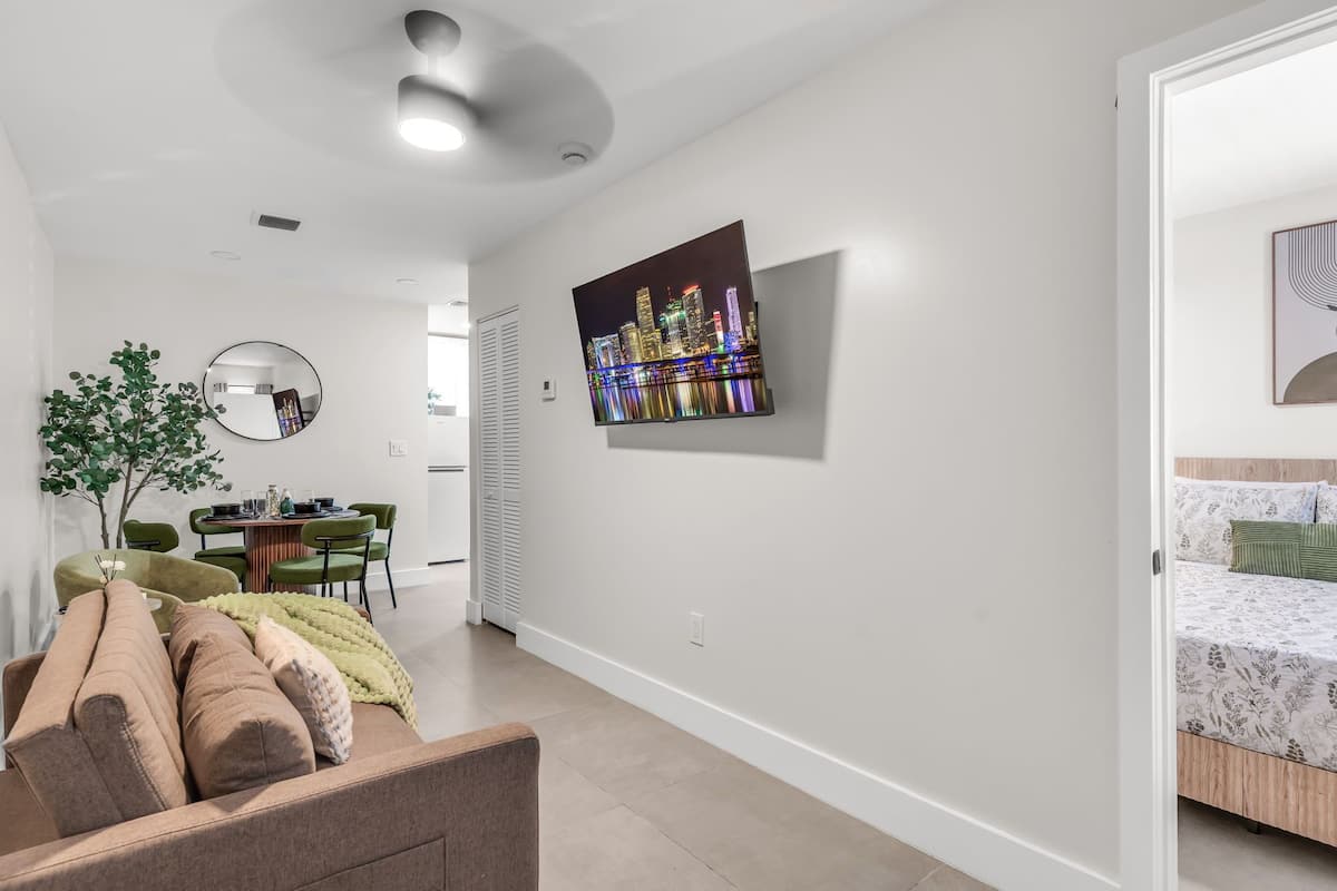 A modern living room with a sofa, a dining area with chairs, a mirror, and a flat-screen TV mounted on the wall.