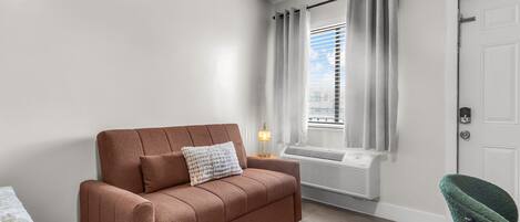 A room with a brown sofa, a lamp, a window with curtains, an air conditioning unit, and a door.