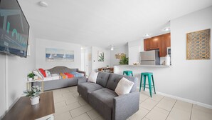 A modern living room with a grey sofa, teal stools, and a kitchen area with wooden cabinets and a stainless steel refrigerator. - Bayview Apartments by Lowkl (Fort Lauderdale)