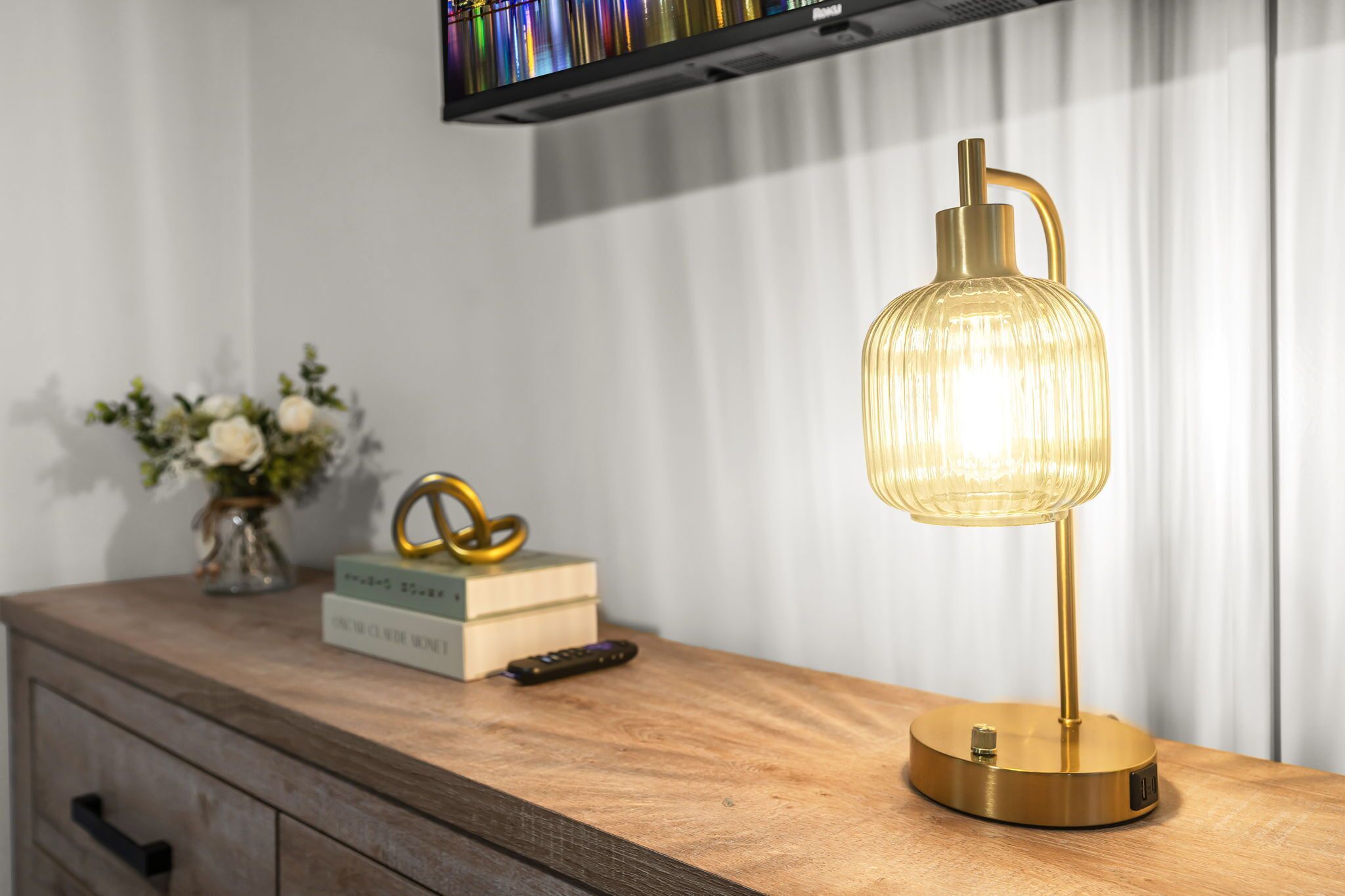 A bedside table with a lamp, a remote control, books, and a vase with flowers.