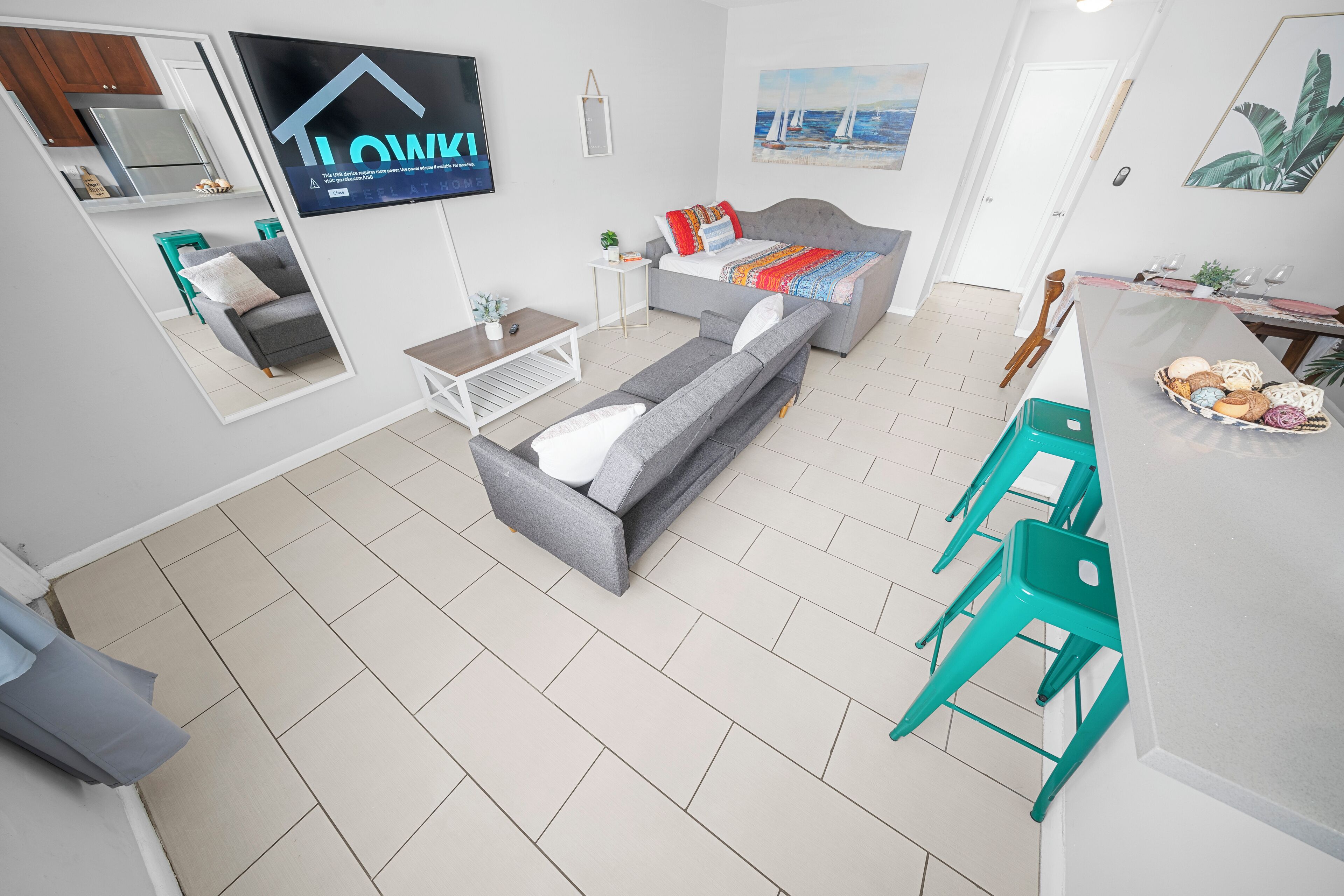 A modern living room with a grey sofa, a flat-screen TV, a dining area with a table and chairs, and a kitchen area with cabinets and a refrigerator.