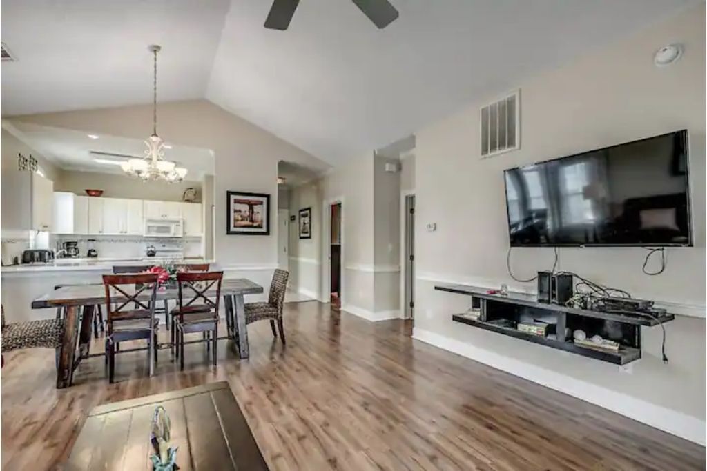 A spacious living area with a dining table, chairs, a flat-screen TV mounted on the wall, and a kitchen in the background.