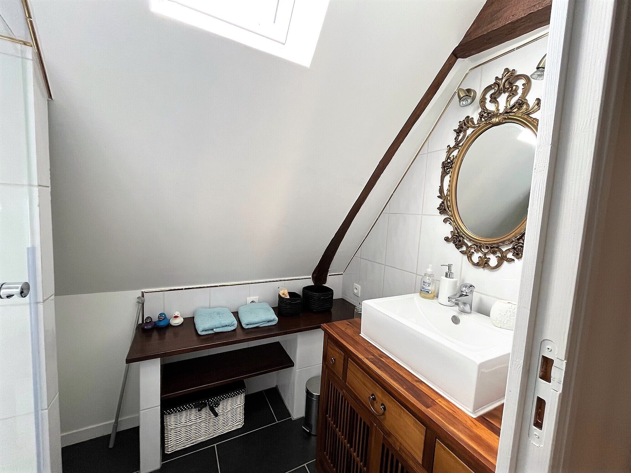 Een compacte badkamer met een witte wastafel, een houten wastafelmeubel, een spiegel met een decoratief kader en een dakraam.