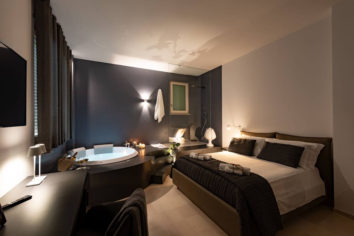 A modern hotel room with a large bed, a round bathtub, a desk with a lamp, and a mirror.