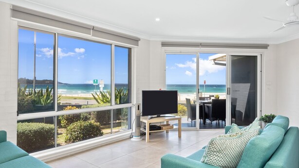 A modern living room with large windows overlooking a beach, a flat-screen TV, and a teal sofa. - Mariners 3 - The closet apartment to Mollymook Beach, you can almost touch it! (Mollymook)