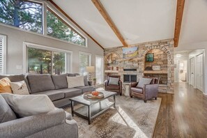 A spacious living room with a stone fireplace, large windows, and a high ceiling with exposed beams.