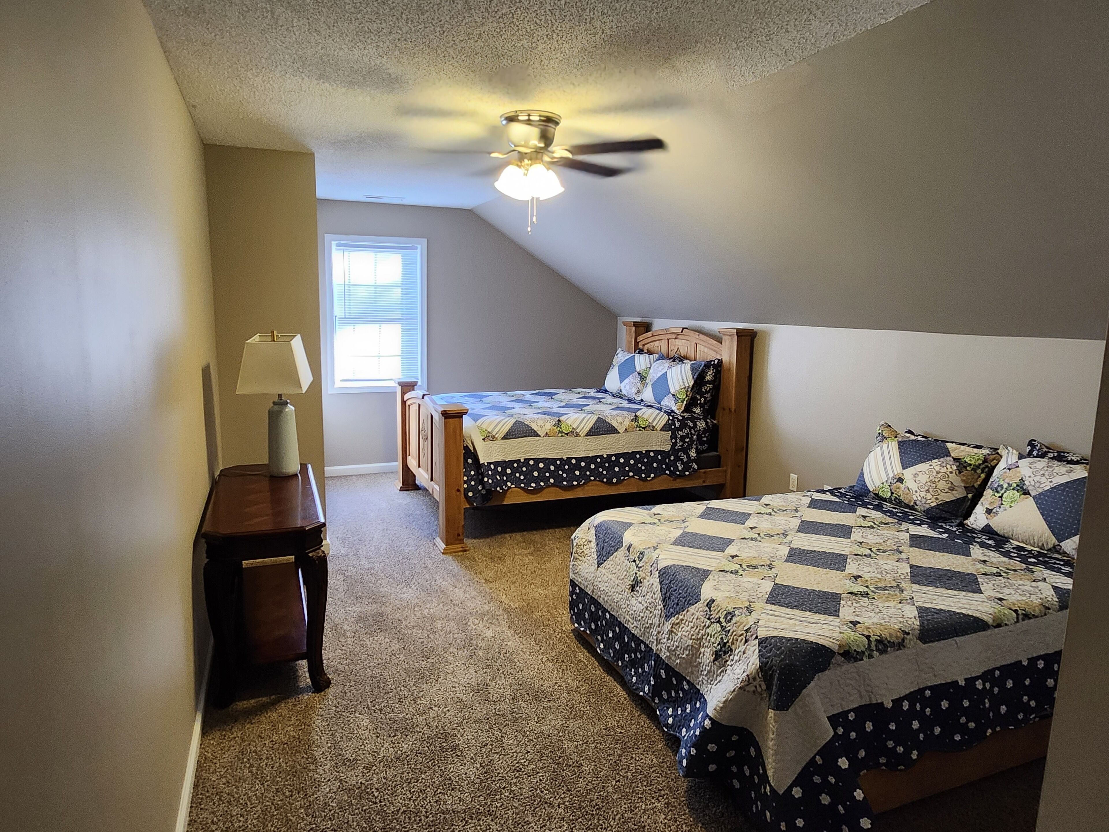 A bedroom with two beds, a ceiling fan, a window, and a nightstand with a lamp.