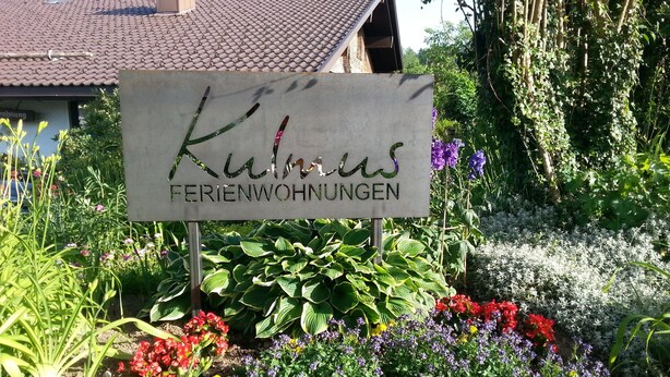 Een bordje voor 'Klinus Ferienwohnungen' omringd door kleurrijke bloemen en begroeiing.