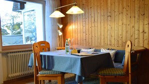 Een eetgedeelte met een tafel voor twee personen, houten stoelen, een blauwe tafelkleed en een raam met uitzicht op bomen.