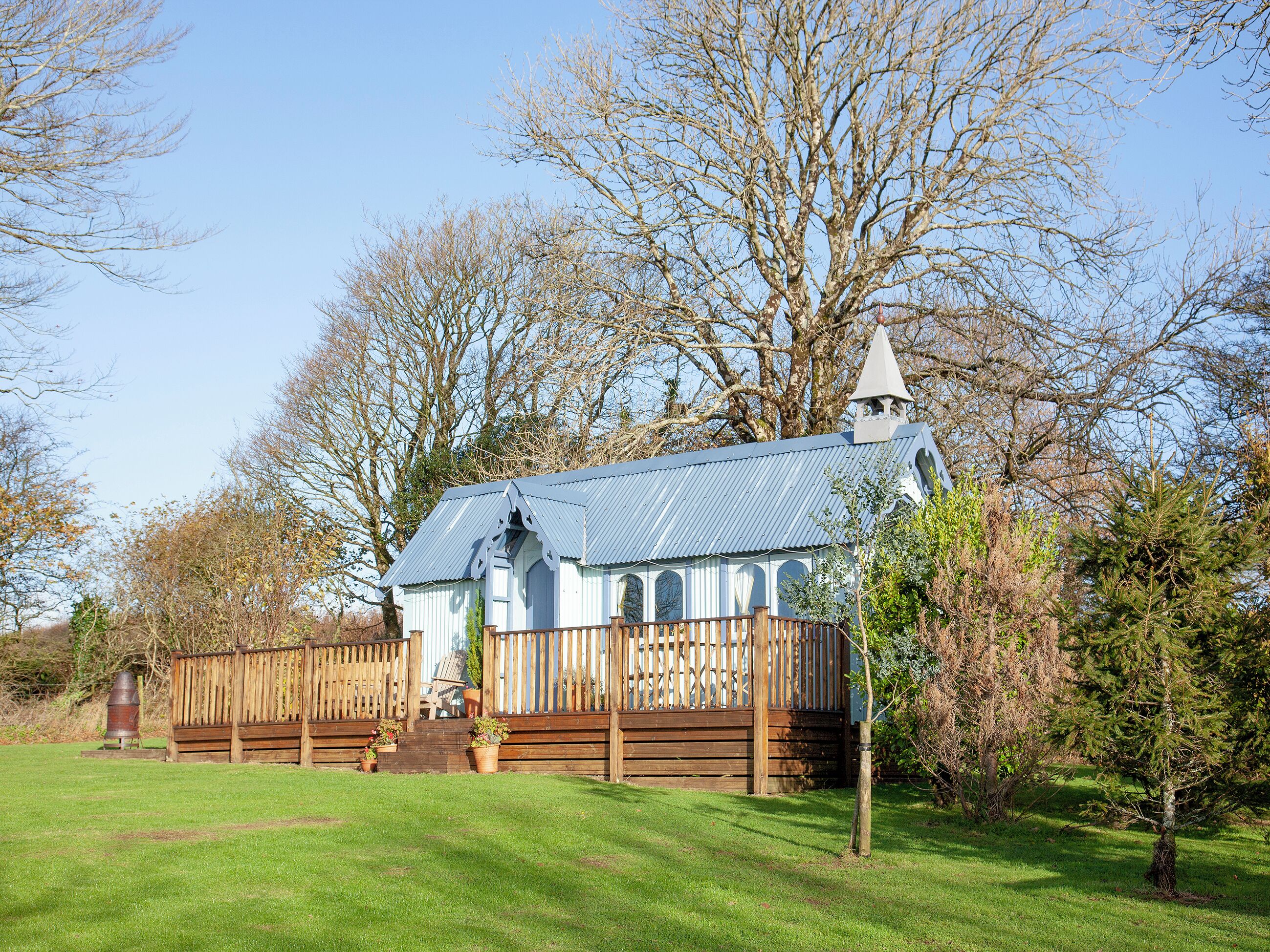 A small, rustic building with a blue roof and a white steeple, surrounded by trees and a wooden fence.