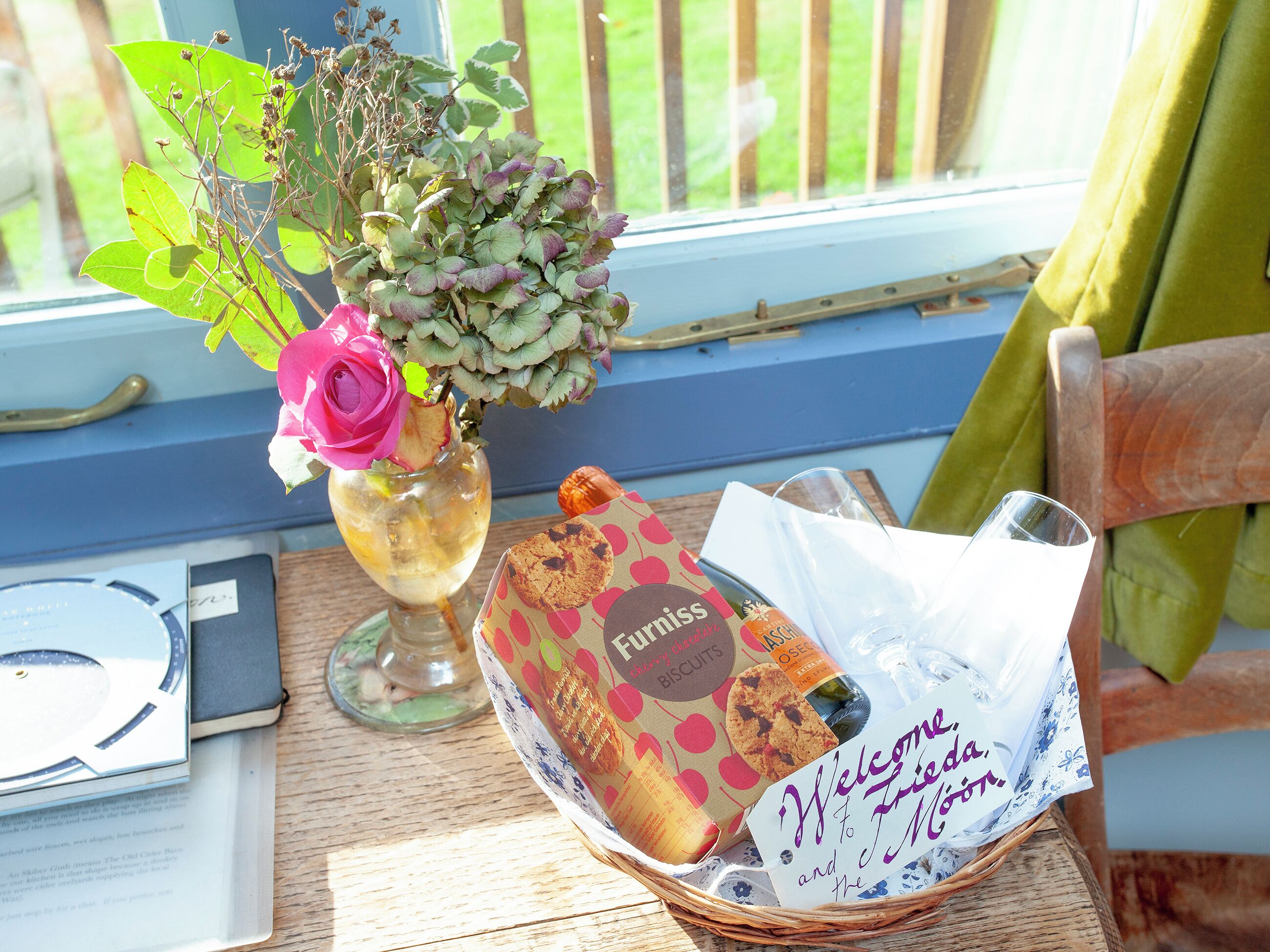 A wicker basket with a welcome note, a cookie, and a bottle of water on a wooden table by a window.