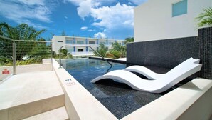 Ein moderner Poolbereich mit einer weißen Liege, Stufen zum Pool und Blick auf ein Hotelgebäude im Hintergrund.