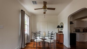 Comedor con mesa y sillas de vidrio, ventilador de techo y vista a una cocina con armarios blancos.