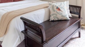 Un dormitorio con una cama, un banco de madera, un ventilador de techo, un cuadro y una ventana con cortinas.