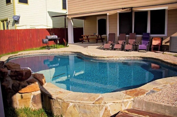 A swimming pool with a stone border, a wooden deck, and a covered patio area with chairs and a table.
