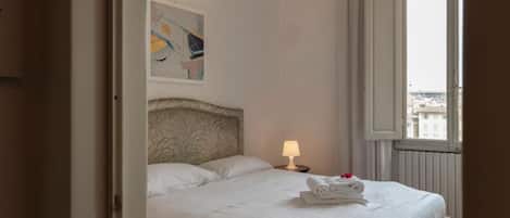 A neatly made bed with white linens, a headboard with a leaf pattern, a bedside lamp, and a window with a view of buildings.
