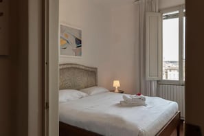 A neatly made bed with white linens, a headboard with a leaf pattern, a bedside lamp, and a window with a view of buildings. - Welcome to Top of Barbadori Tower – River View Apartment by Ponte Vecchio, a unique stay inside one of Florence’s historic tower buildings. Stay in a bright top-floor apartment inside an authentic 12th-century Florentine tower, just steps from Ponte  (Firenze)