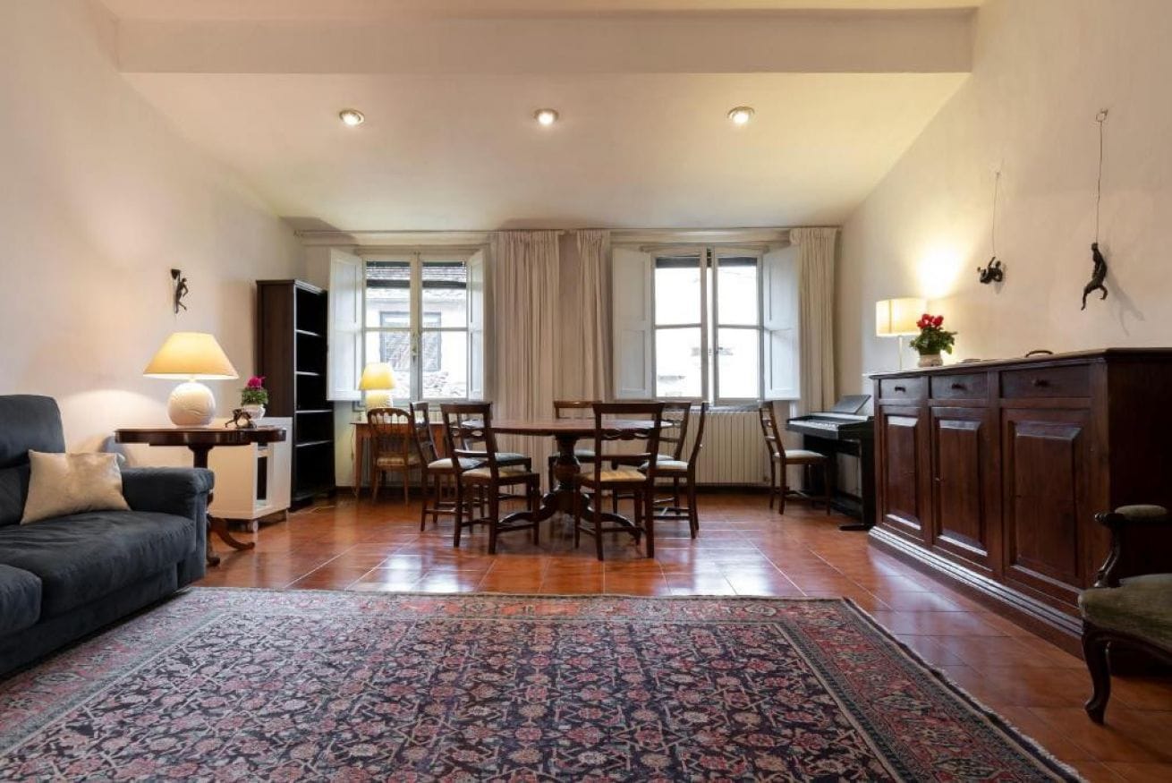 A spacious living area with a dark-colored sofa, a patterned rug, a dining table with chairs, and a kitchen area with cabinets and a countertop.