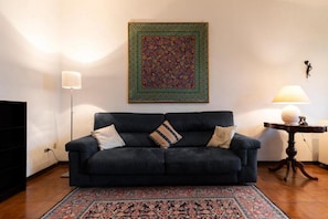 A living room with a dark sofa, patterned rug, and a decorative wall hanging. - Welcome to Top of Barbadori Tower – River View Apartment by Ponte Vecchio, a unique stay inside one of Florence’s historic tower buildings. Stay in a bright top-floor apartment inside an authentic 12th-century Florentine tower, just steps from Ponte  (Firenze)