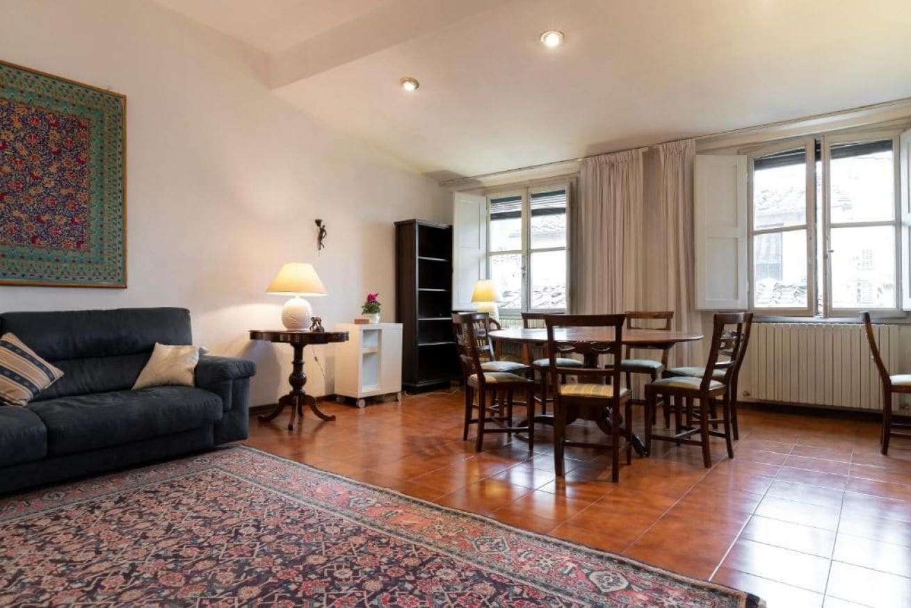 A living room with a dark sofa, a patterned rug, a dining table with chairs, and a bookshelf.
