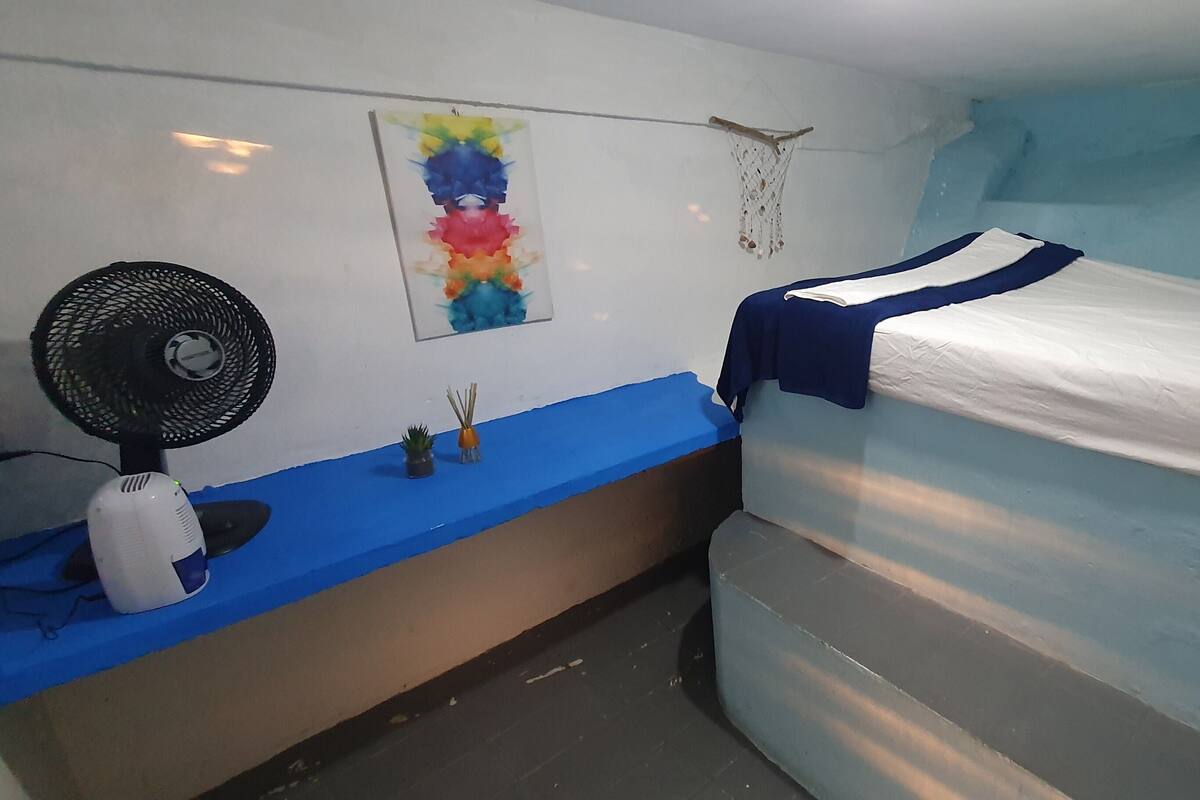Une petite pièce, de taille modeste, comprenant un lit simple, une table de chevet, un ventilateur fixé au mur et un tableau aux couleurs vives.