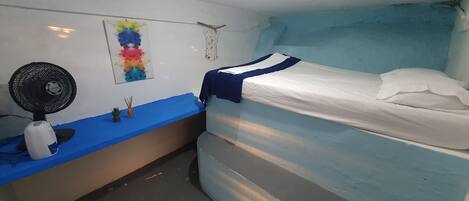 A small, compact room with a single bed, a bedside table, a wall-mounted fan, and a colorful painting.