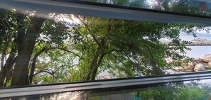 A view through a window with trees and a body of water in the background. - Hostel Farol da Barra (Florianópolis)