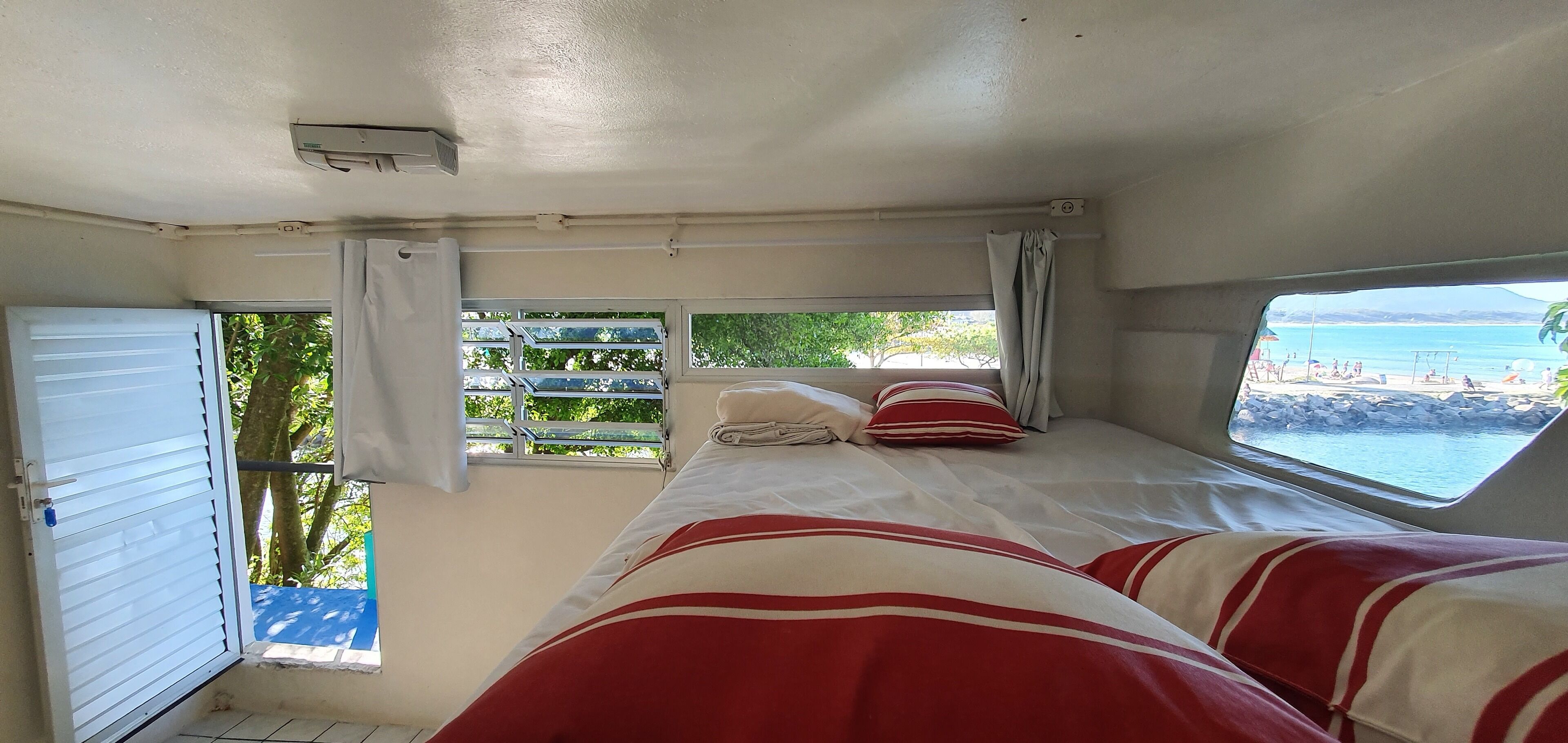 A room with a bed, a window with a view of a beach, and a door.