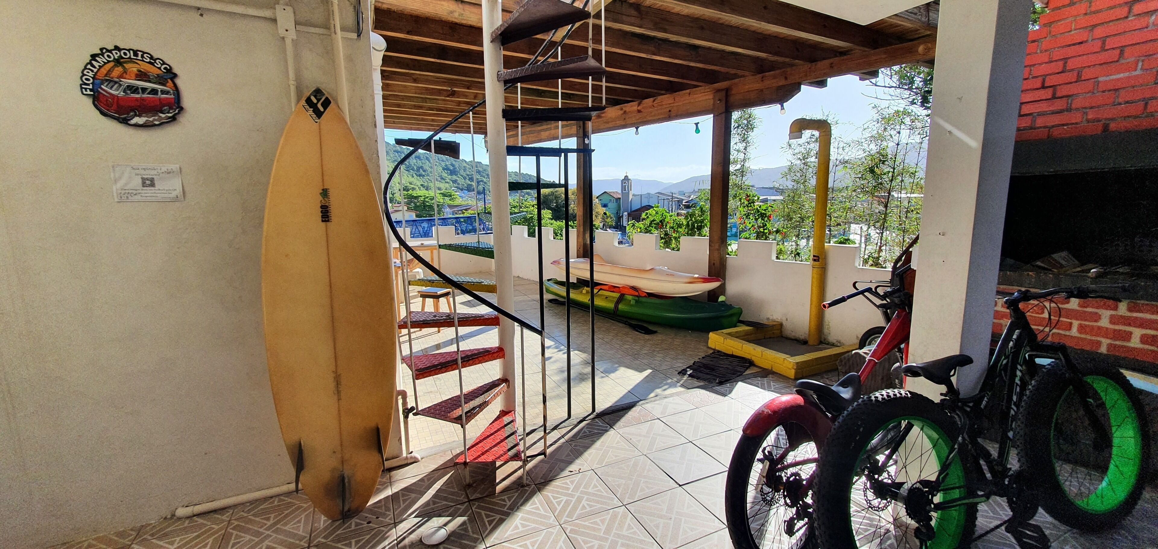 A covered outdoor area with a surfboard leaning against the wall, a bicycle, and a staircase leading to a terrace with a view of the hills.
