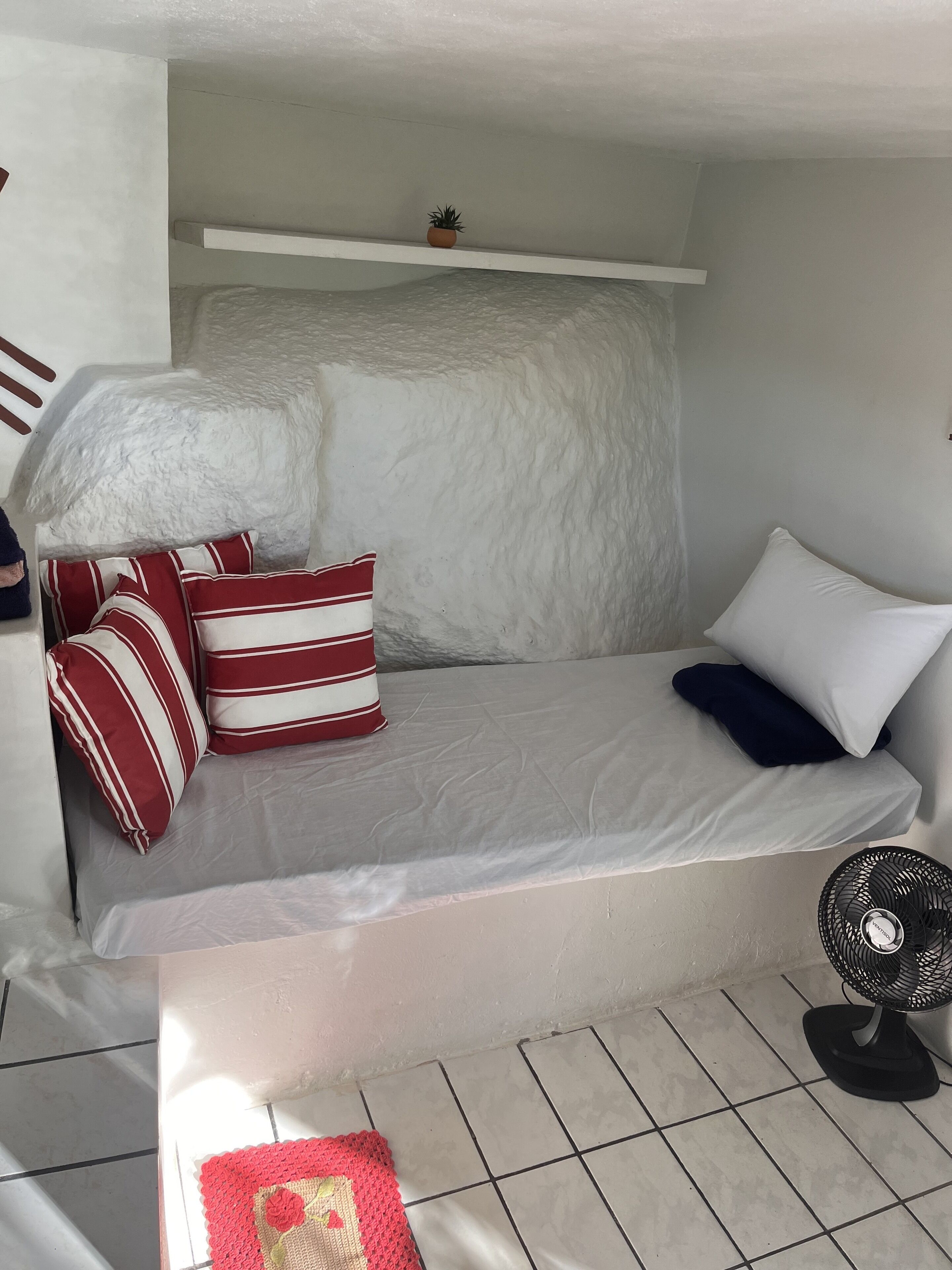 A small, compact room with a bed, pillows, a fan, and a small potted plant.