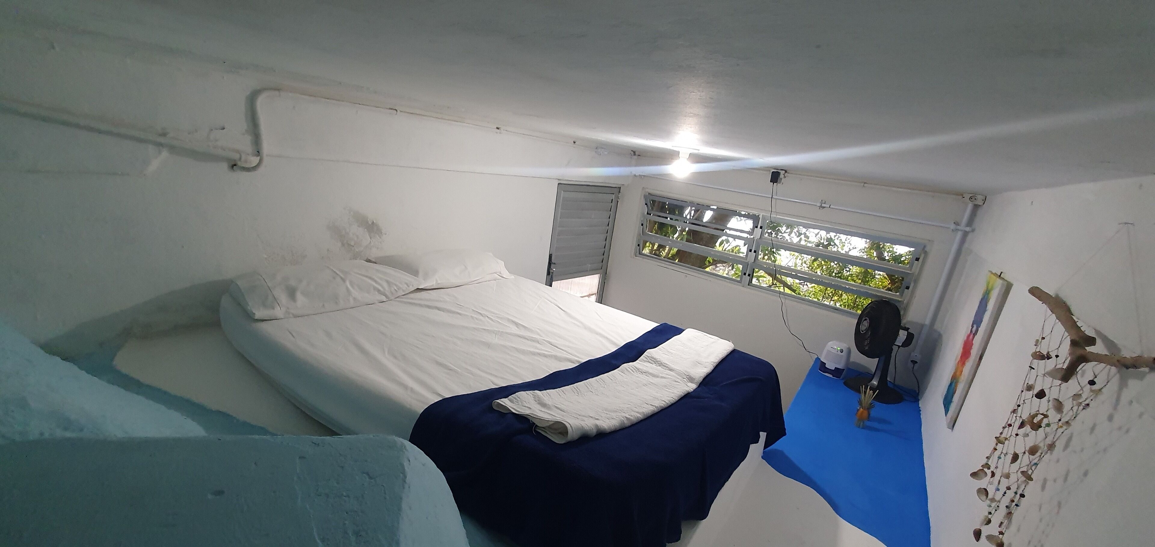 A small, simple room with a bed, a window with blinds, a small table with a fan, and a wall decoration.