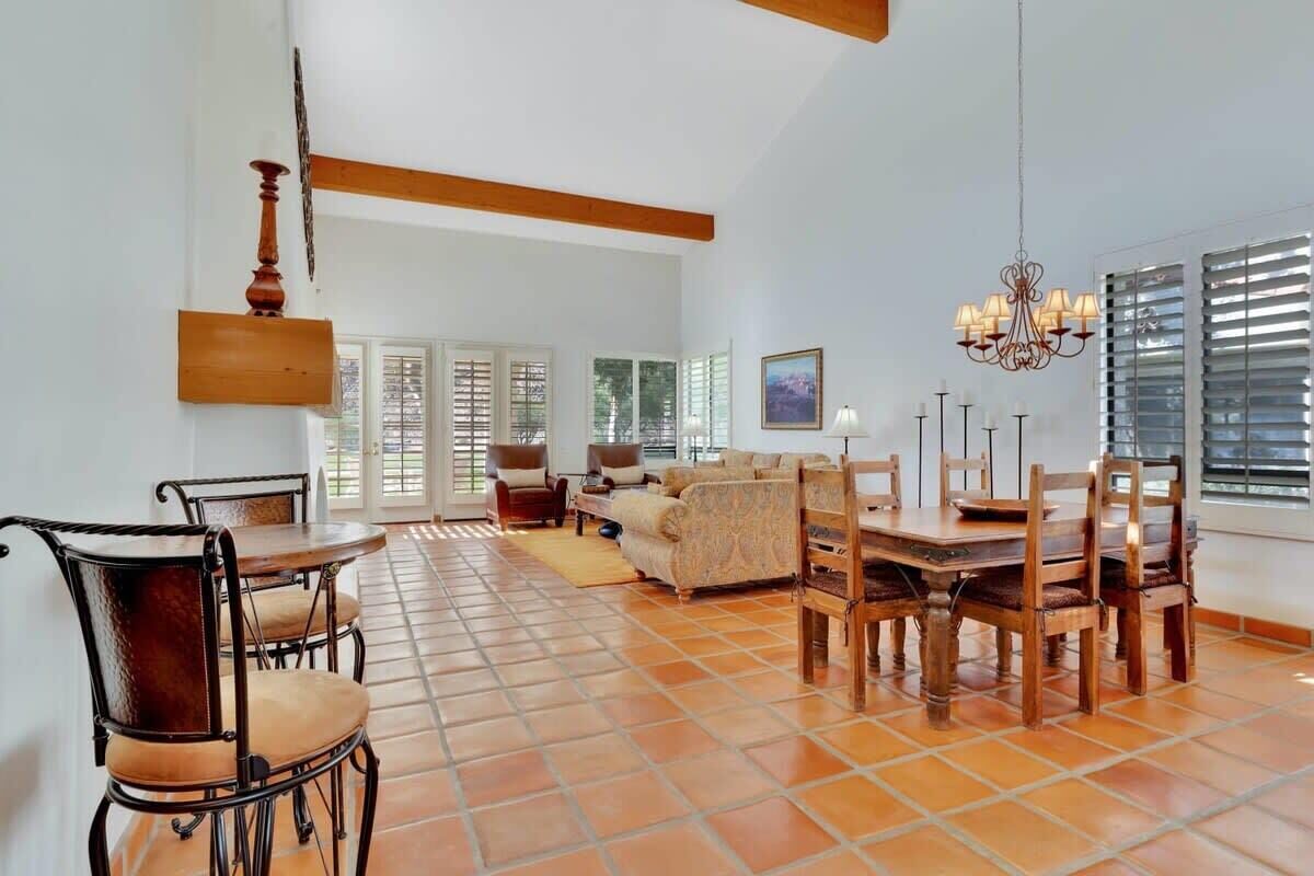A spacious living room with a dining area, featuring a tiled floor, wooden furniture, and large windows with blinds.