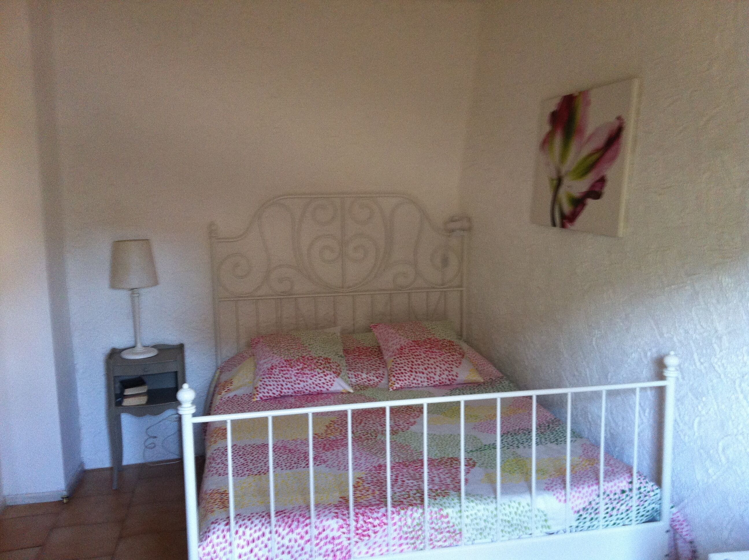 Ein Schlafzimmer mit einem Bettgestell aus weißem Metall, einem Nachttischchen mit Lampe und einem floralen Wandbild.