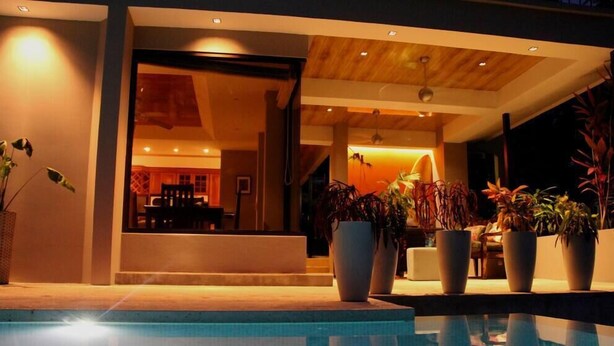 A modern pool area with a clear view of the pool, potted plants, and a glimpse of the interior space with a wooden ceiling and warm lighting. - Moonshine Inn — Pool Level Suite , Moonshine Inn -- Pool-Level 2BR Suite (Quepos)