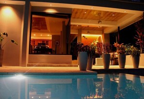 Ett modernt poolområde med fri sikt över poolen, krukväxter och en inblick i det inre utrymmet med ett träbeklätt tak och behaglig belysning.