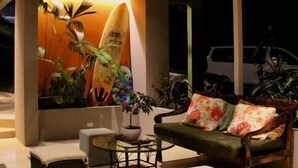 A modern living room with a wooden ceiling, a ceiling fan, a surfboard mural, a sofa with floral cushions, a small table, and a potted plant. - Moonshine Inn — Pool Level Suite , Moonshine Inn -- Pool-Level 2BR Suite (Quepos)