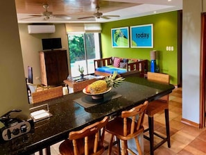 A kitchen with a central island, wooden chairs, a pineapple on the island, and a television on the wall.