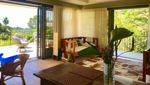 A room with a bed, a table, chairs, a ceiling fan, and large windows with curtains. - Moonshine Inn - 1 BR Studio Loft with Ocean & Jungle Views, Moonshine Inn -- 1BR Studio Loft (Quepos)