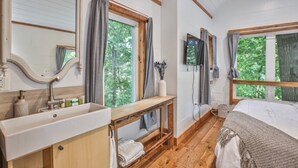 A bedroom with a wooden floor, a bed, a bedside table, a mirror, and a window with curtains.