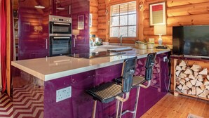 A wooden cabin interior with a kitchen area featuring a purple island, two ovens, a sink, and a television. - Grebe Lodge (Lodge 6) (Frettenham)