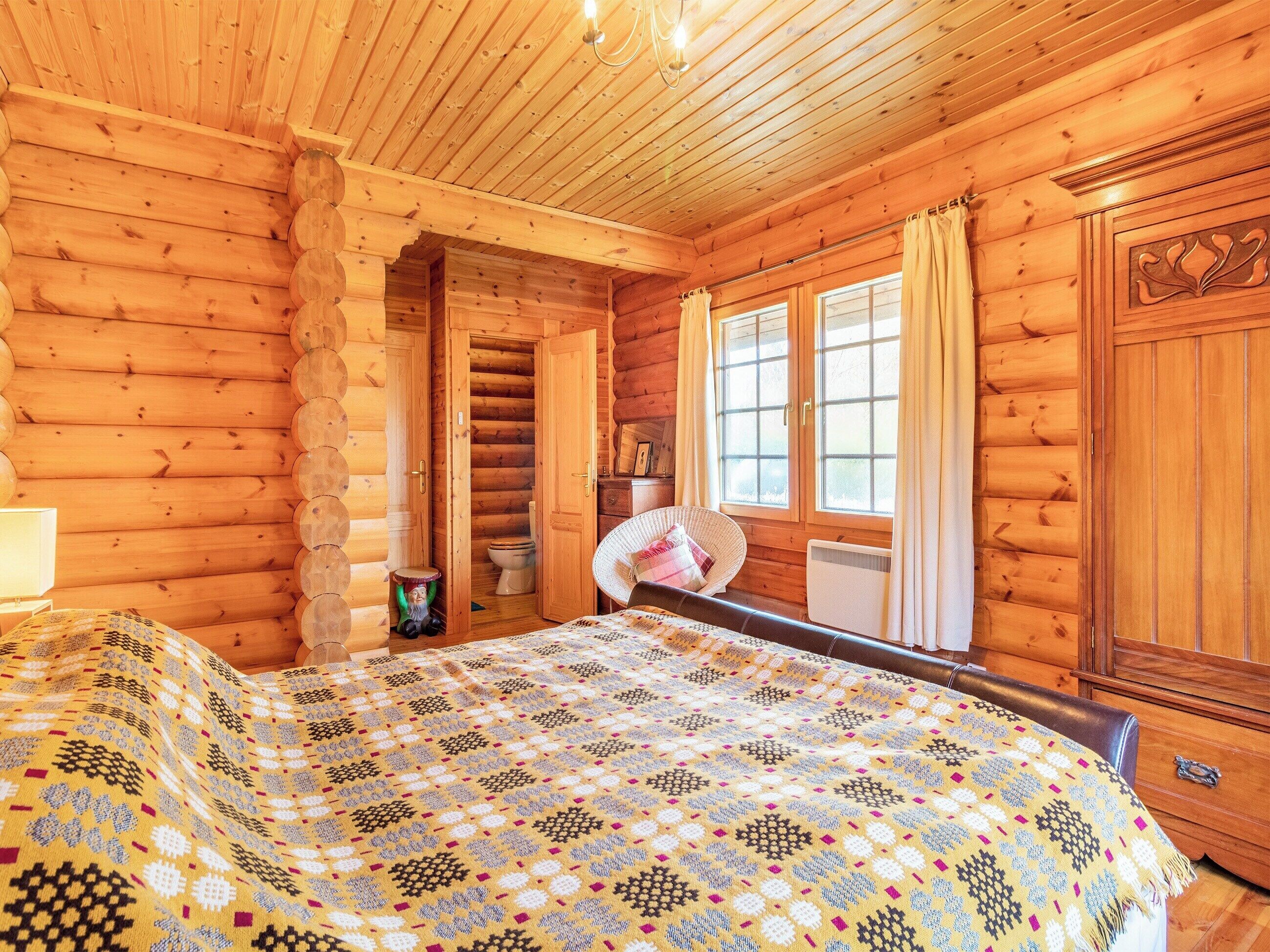 Habitació d'una cabana de fusta amb un llit, una finestra amb cortines, un armari de fusta i un petit tamboret.