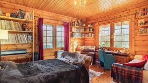 A cozy log cabin room with a bed, a desk, and a chair. There are bookshelves filled with books and a window with curtains. - Grebe Lodge (Lodge 6) (Frettenham)