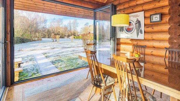 A wooden cabin interior with a dining area, a large window, and a poster on the wall. - Grebe Lodge (Lodge 6) (Frettenham)