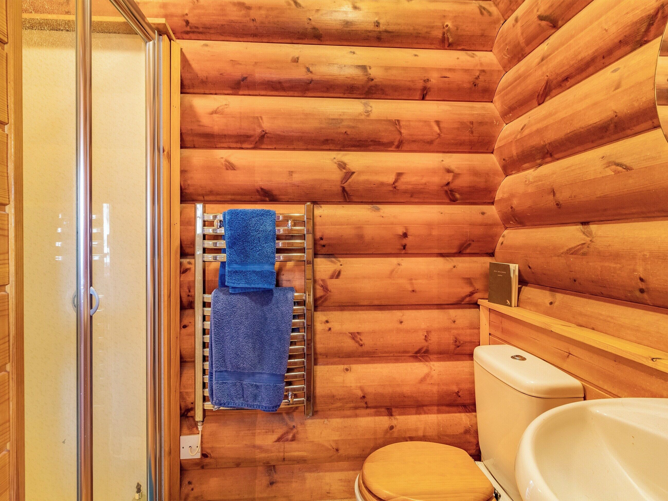 Une salle de bain rustique, avec des murs en bois, comprenant des toilettes, un porte-serviettes avec deux serviettes et une cabine de douche en verre.