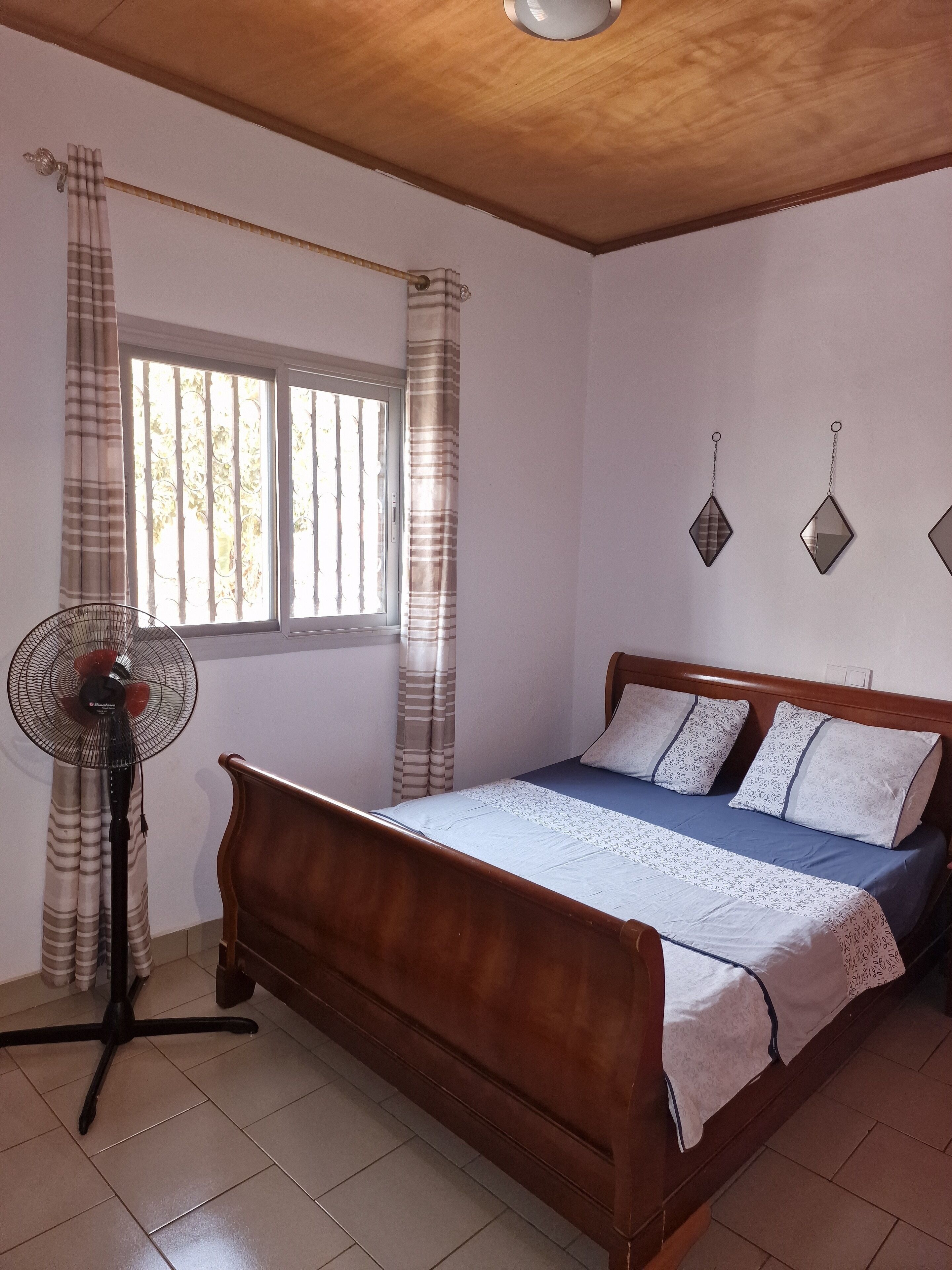 Un dormitorio con cama de madera, un ventilador de pie y una ventana con cortinas.