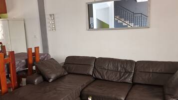Ein Wohnzimmer mit einem braunen Ledersofa, einem kleinen runden Tisch mit einer Vase und einem weiteren runden Tisch mit einem gelben Topf. Ein Spiegel hängt an der Wand und spiegelt eine Treppe wider.